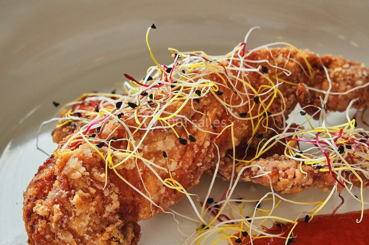 Plat de langoustine panée garnie de pousses colorées, accompagnée d'une sauce rouge servie dans une assiette blanche.