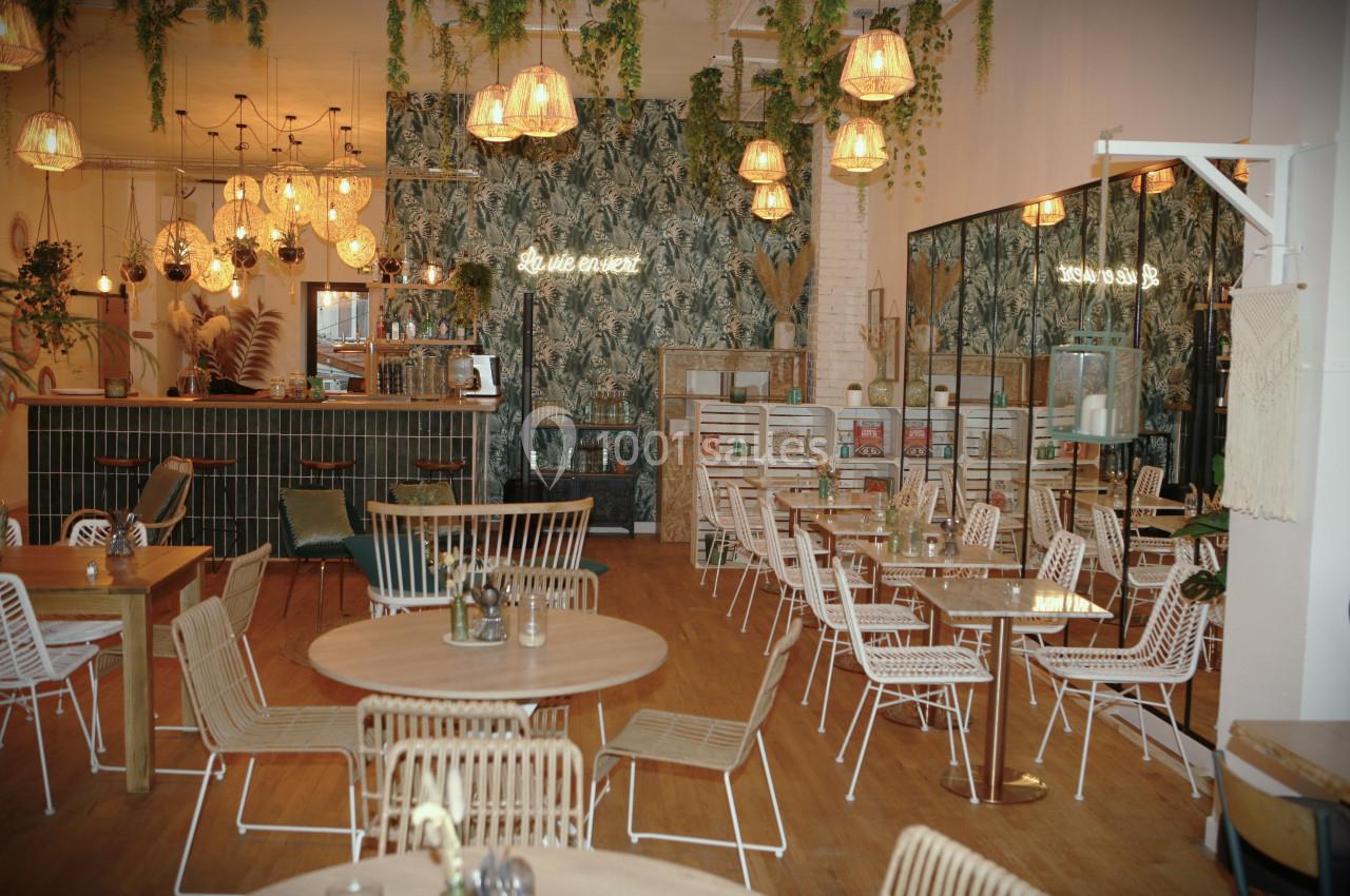 Salle de restaurant lumineuse avec mobilier en rotin, plantes suspendues et décoration murale tropicale.