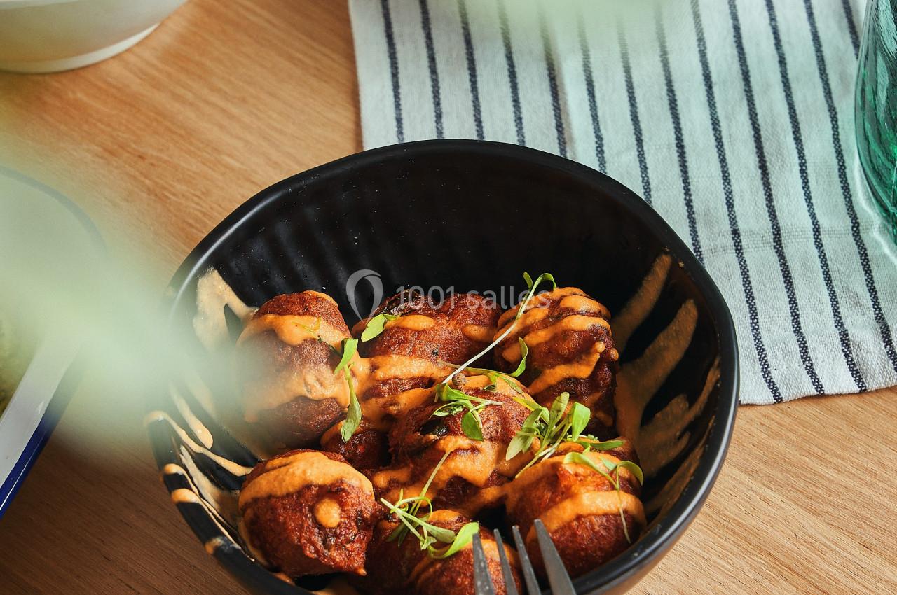 Bol de bouchées dorées garnies de sauce et de micro-pousses, posé sur une table en bois avec des plats en arrière-plan.