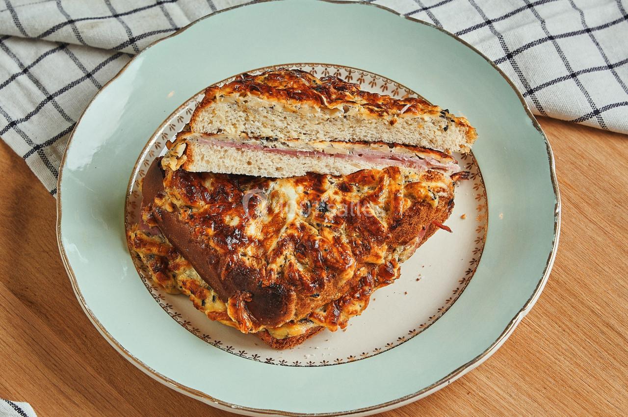 Part de croque-monsieur gratiné sur une assiette blanche bordée de motifs, posée sur une table en bois avec une nappe à…