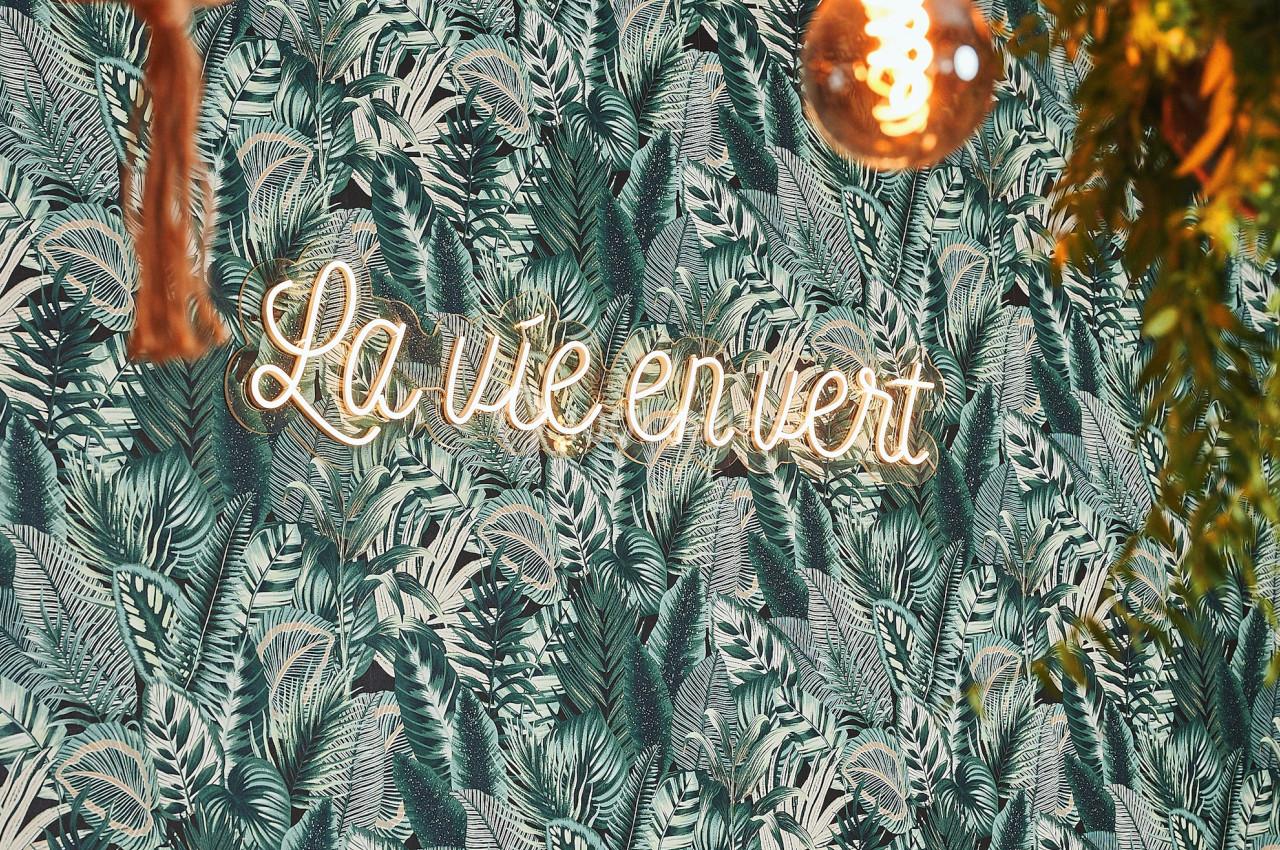 Mur décoré d'un papier peint à motifs végétaux, éclairé par une enseigne néon ’La vie en vert’ et des suspensions lumineuses.