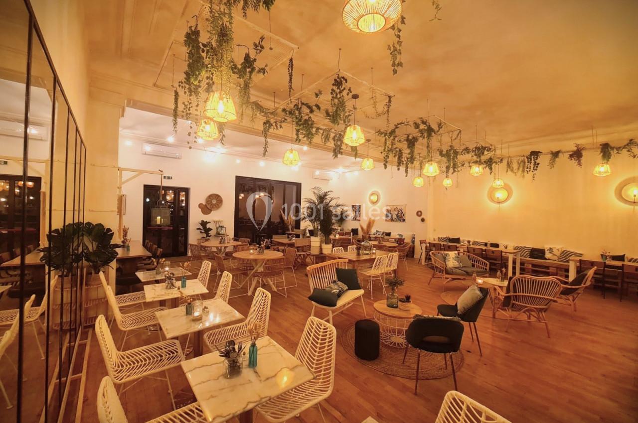 Salle de restaurant chaleureuse avec éclairage tamisé, plantes suspendues et mobilier en bois et métal.
