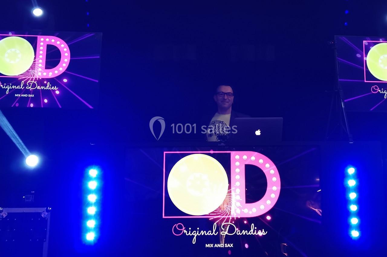 Un DJ mixant sur une scène éclairée avec des écrans lumineux et un saxophoniste à gauche.