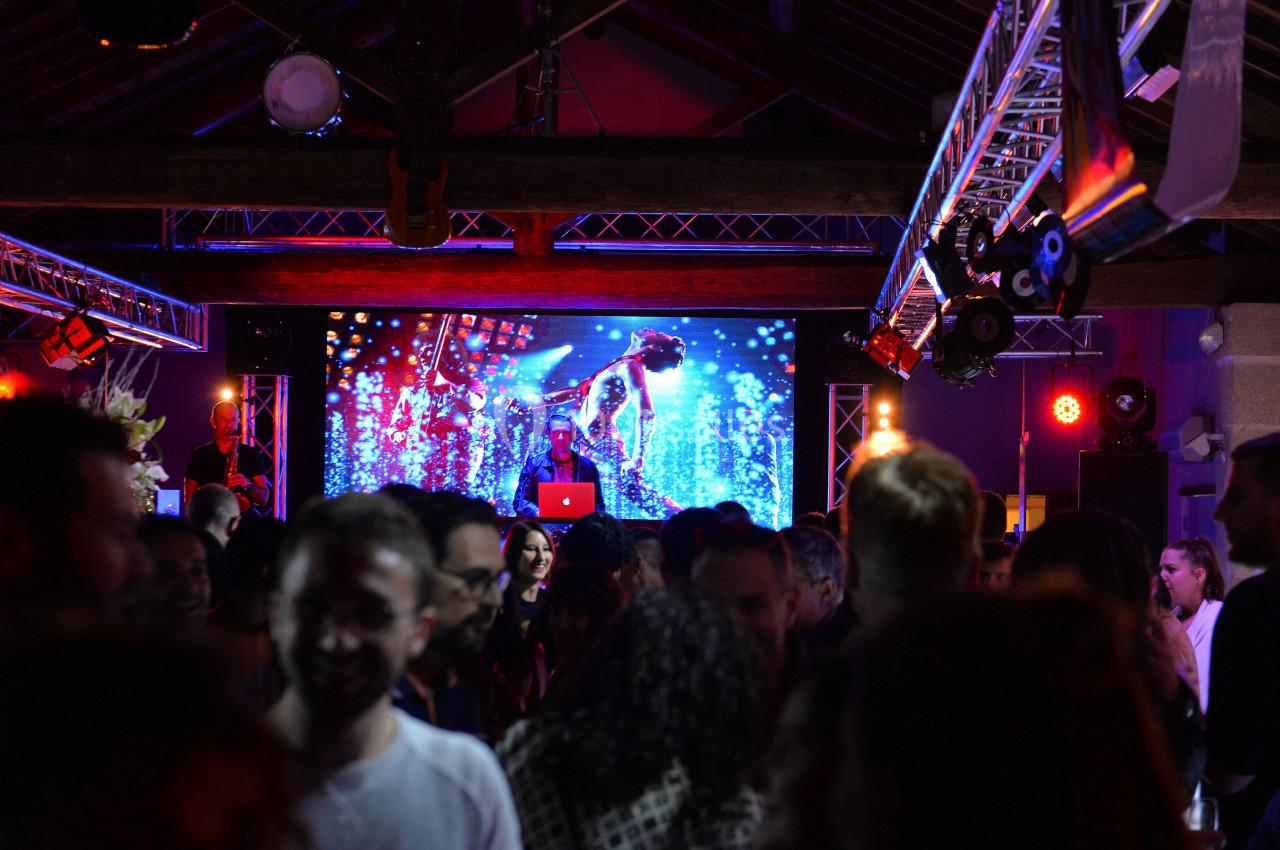 Un DJ mixe sur scène devant un écran lumineux, entouré d'une foule dans une ambiance festive et colorée.