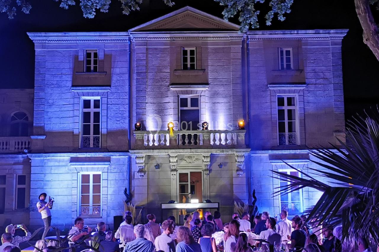 Façade d'un bâtiment éclairé en bleu la nuit, avec des personnes assises devant lors d'un événement en extérieur.