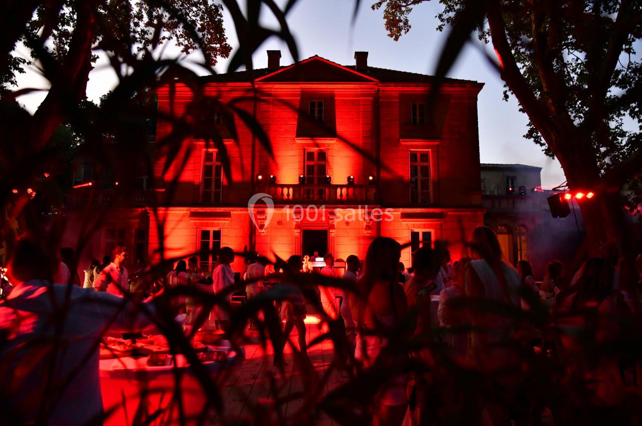 Façade d'un bâtiment éclairé en rouge au crépuscule, entouré d'arbres et d'une foule en extérieur.