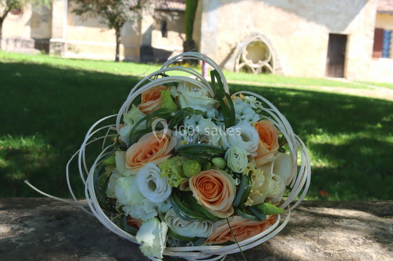 Bouquet de fleurs aux tons orange et blanc posé sur une pierre, avec un jardin et un bâtiment en arrière-plan.
