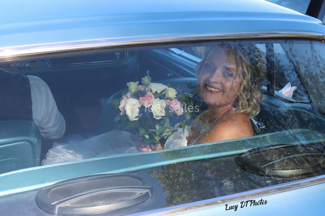 Une femme souriante en robe de mariée tient un bouquet de fleurs dans une voiture ancienne.