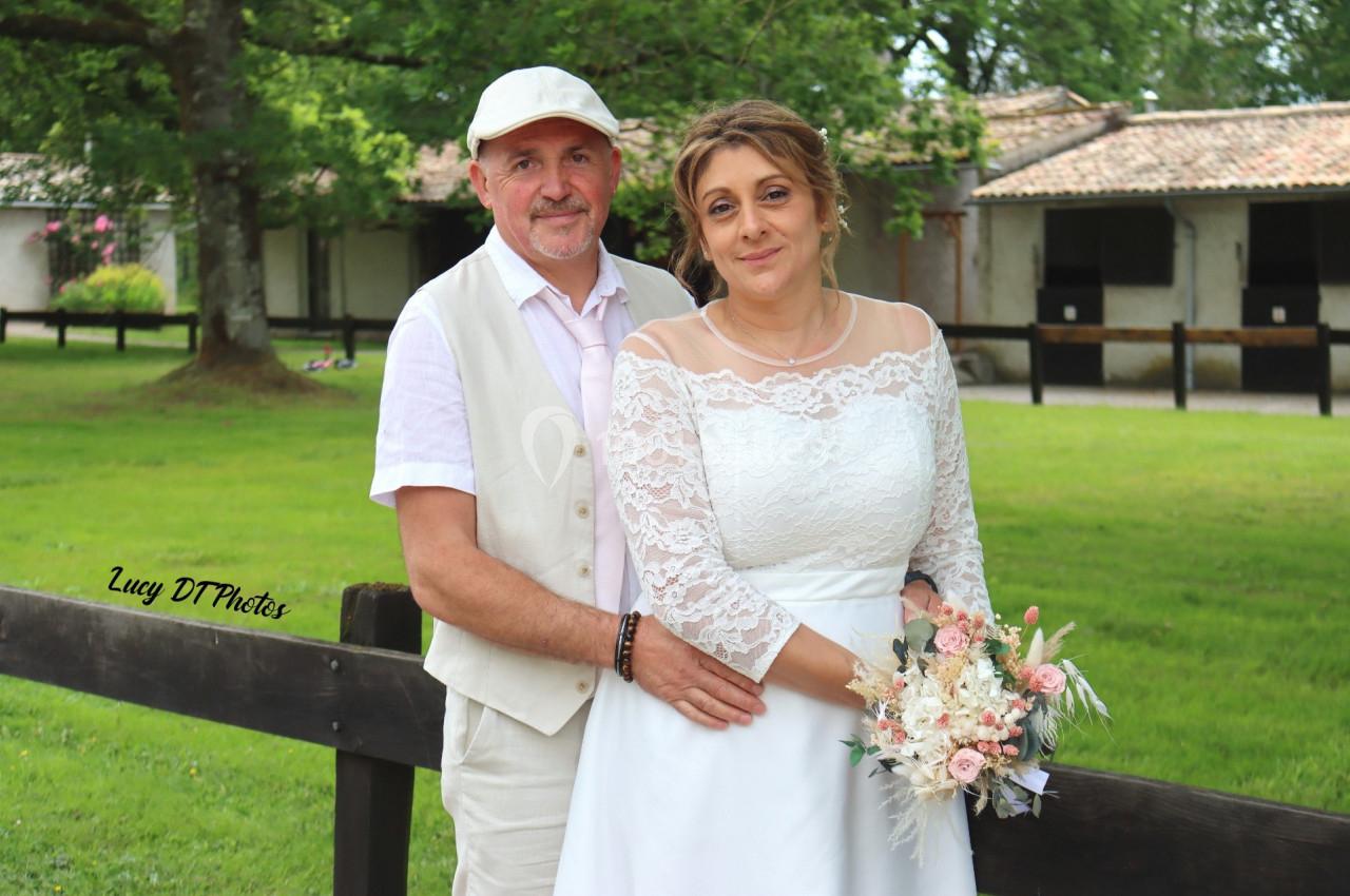 Un couple en tenue de mariage pose devant une clôture en bois, avec des bâtiments et un espace vert en arrière-plan.