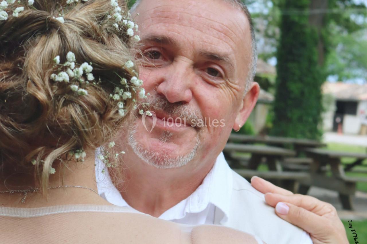 Un homme souriant regarde vers l'objectif, tenant une femme en robe de dentelle blanche dans un parc verdoyant.