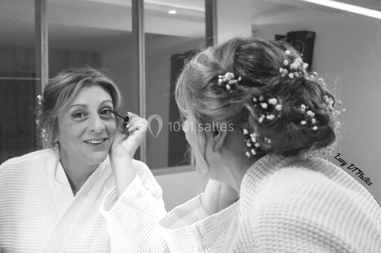 Femme en peignoir blanc se maquillant devant un miroir, ses cheveux ornés de petites fleurs blanches.