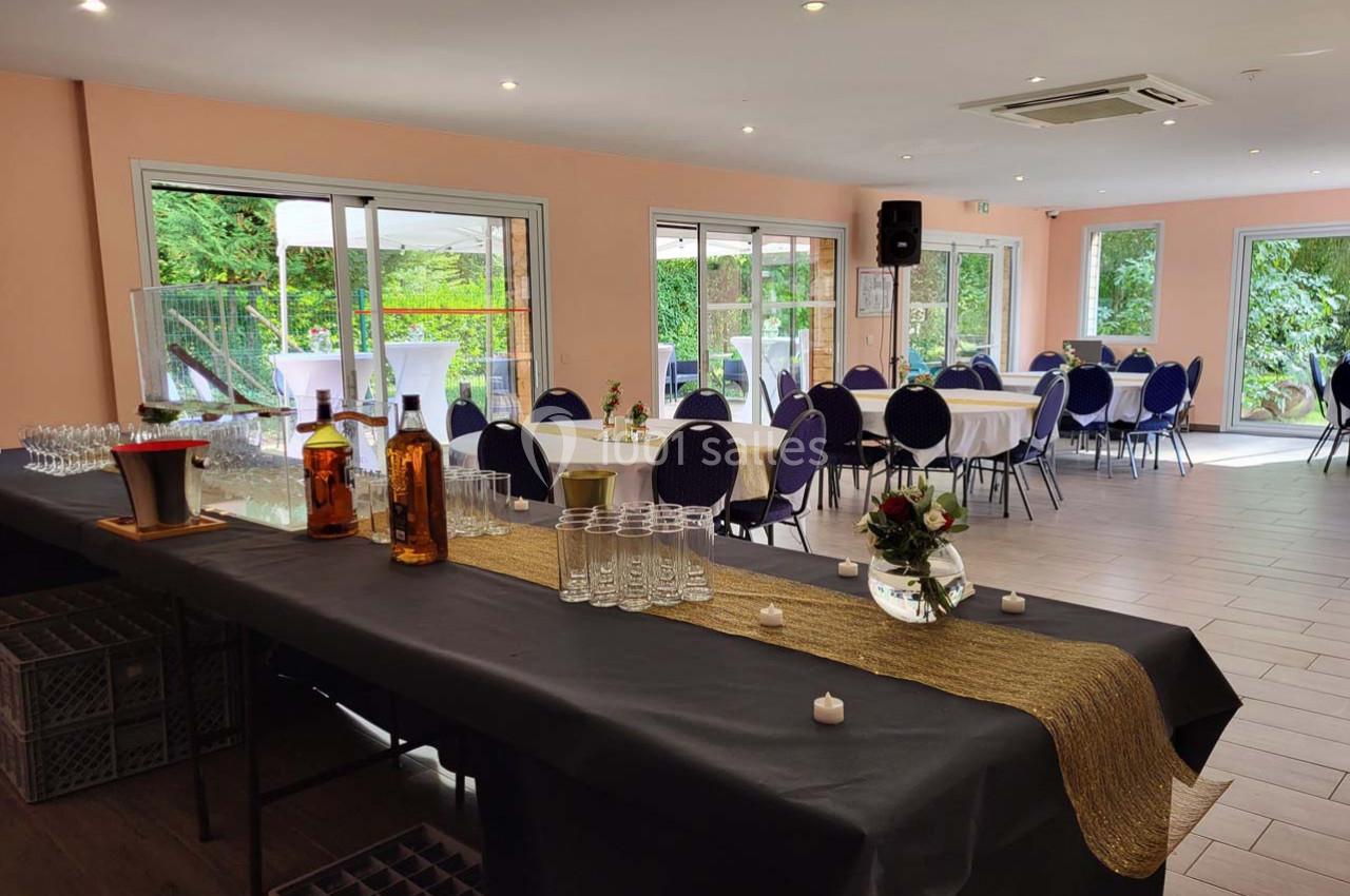 Salle lumineuse avec tables rondes dressées, chaises bleues et buffet décoré, vue sur un jardin à travers de grandes baies…