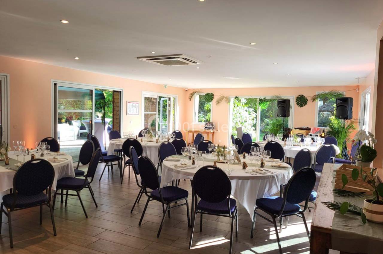 Salle lumineuse aménagée pour un événement, avec des tables rondes dressées et des chaises noires, vue sur un jardin.