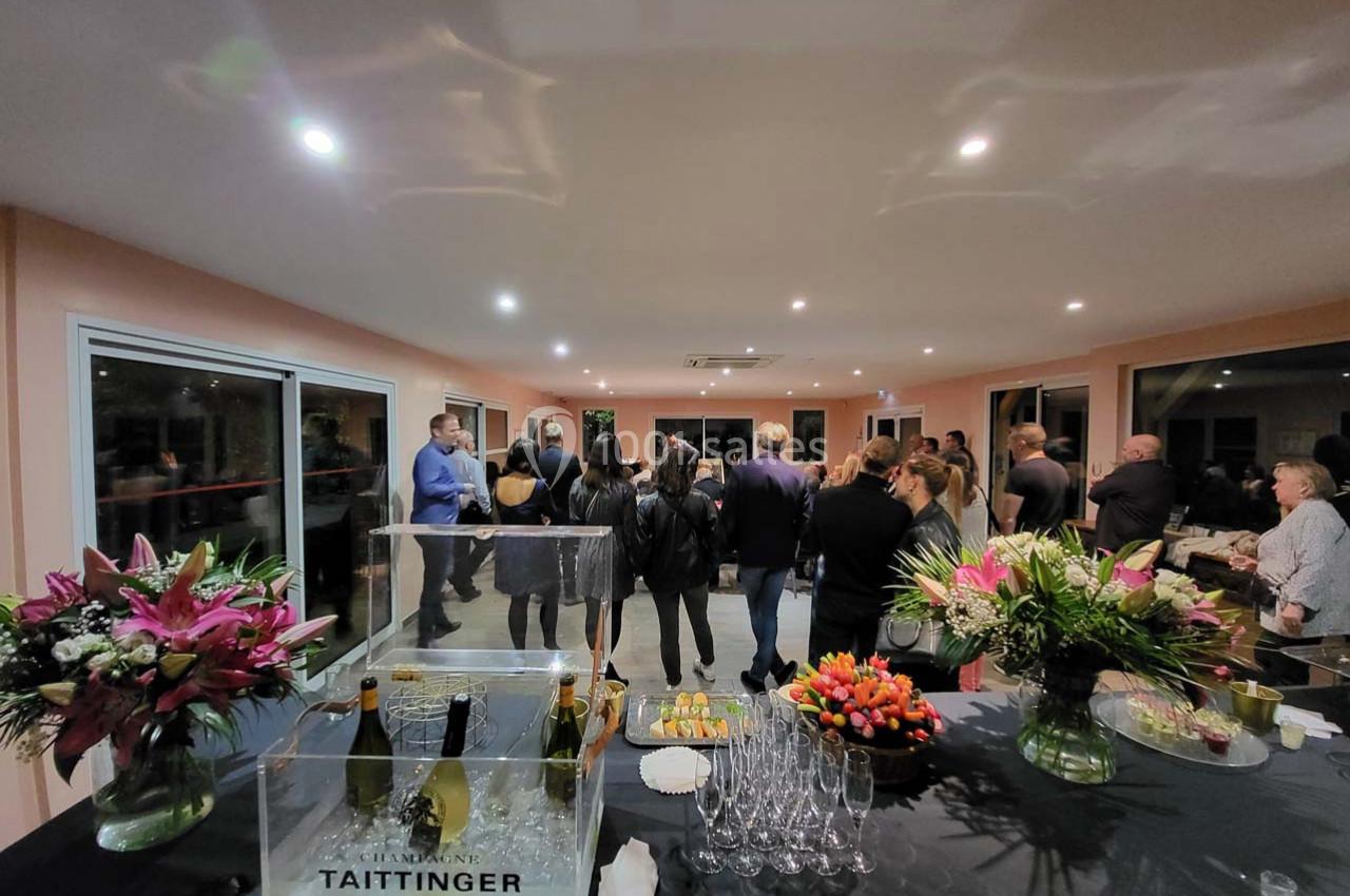 Groupe de personnes rassemblées dans une salle lumineuse, avec une table garnie de fleurs, verres et bouteilles de champagne.