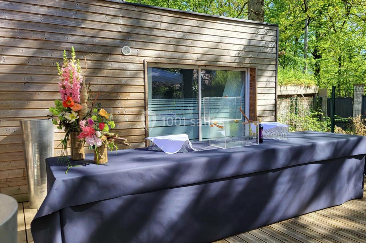Table drapée de tissu bleu avec des décorations florales et une boîte transparente, située sur une terrasse en bois.