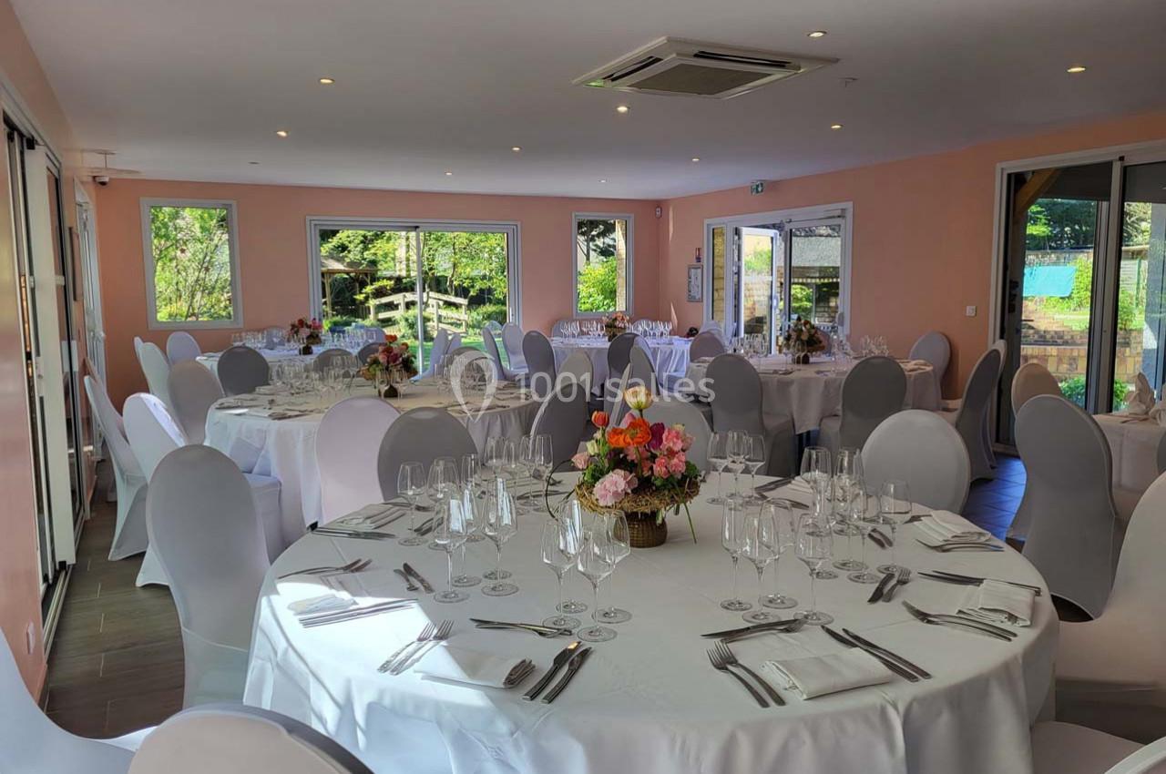Salle de réception lumineuse avec tables rondes dressées, nappes blanches et compositions florales au centre.