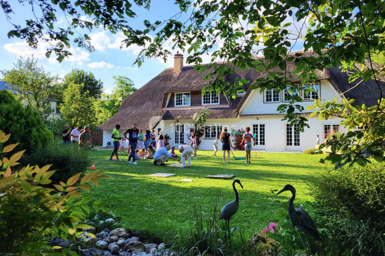 Un groupe de personnes joue et discute dans le jardin d'une maison à toit de chaume, entourée de verdure.