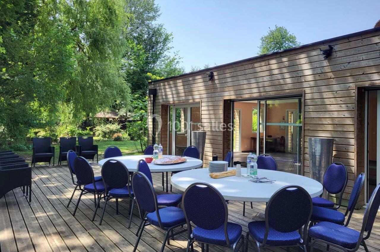 Terrasse en bois avec tables rondes dressées, chaises bleues et bâtiment en bois entouré de verdure.