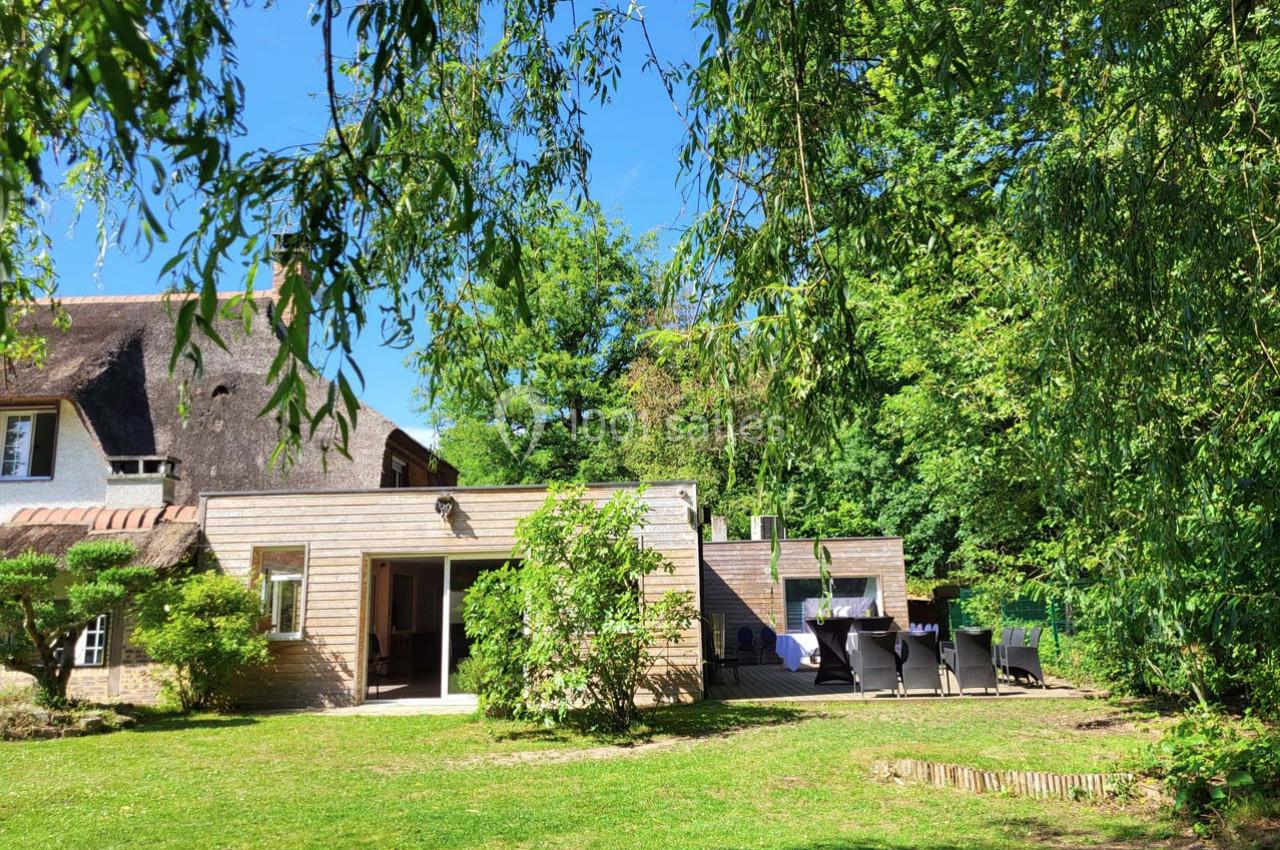 Maison avec toit en chaume entourée d'un jardin verdoyant, terrasse aménagée avec table et chaises sous un ciel bleu.