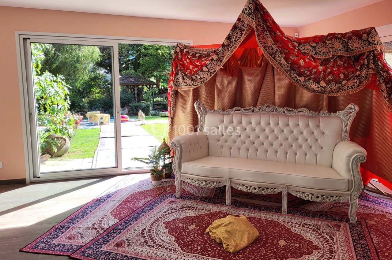 Canapé blanc devant une tente décorative rouge et dorée, posé sur des tapis orientaux, avec vue sur un jardin ensoleillé.