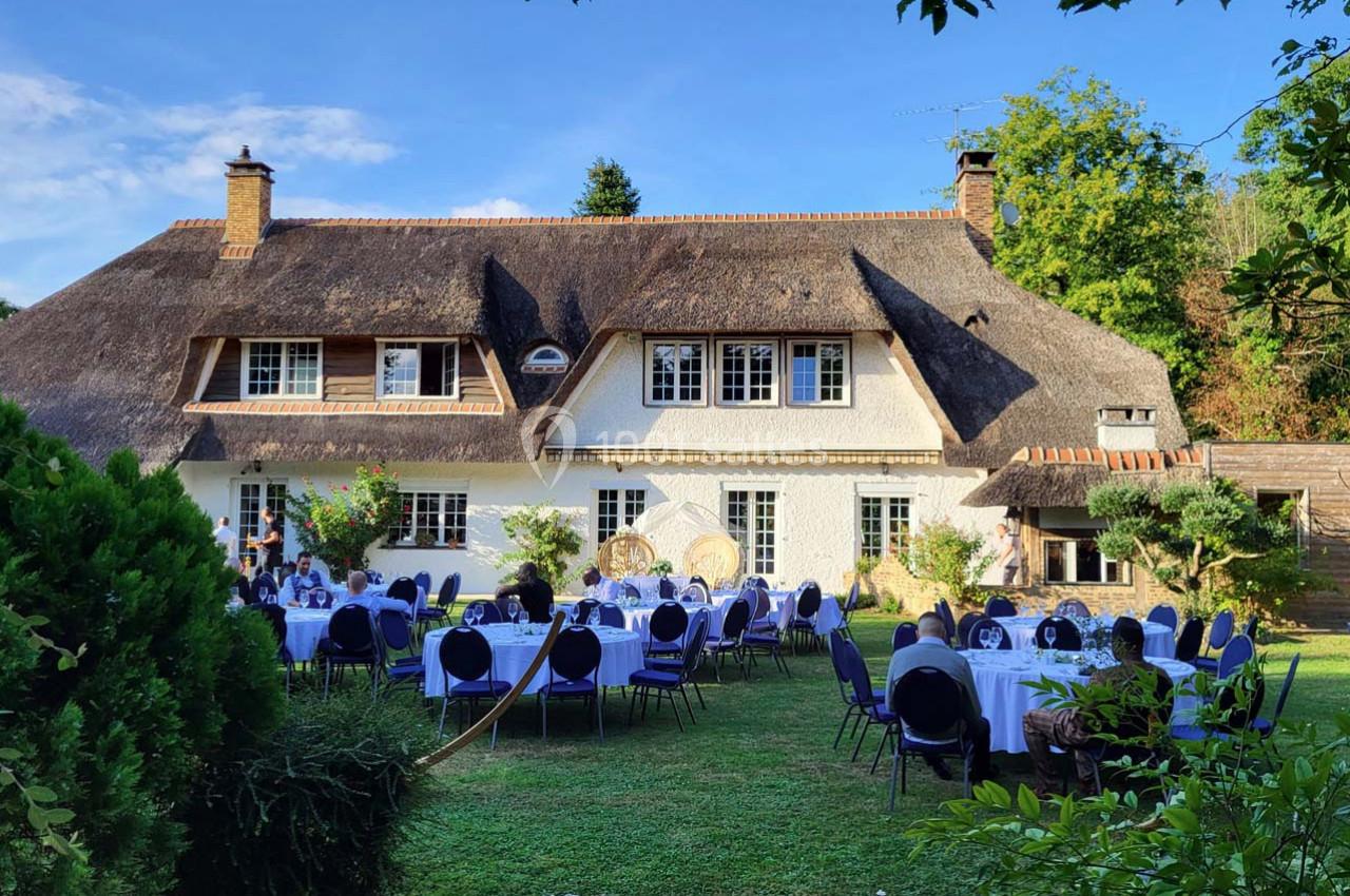 Tables dressées dans un jardin verdoyant devant une maison à toit de chaume, sous un ciel bleu.