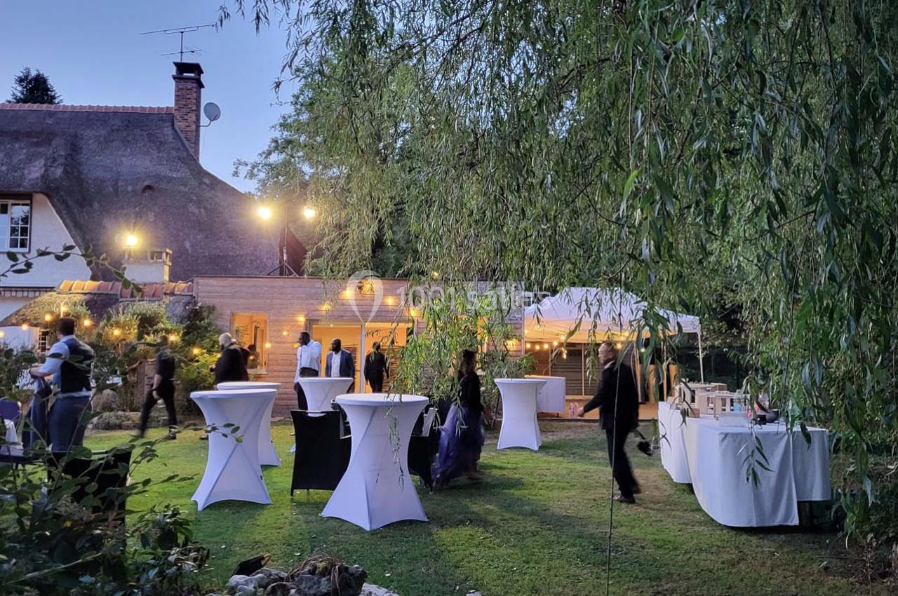 Jardin éclairé en soirée avec des tables hautes blanches et des invités debout près d'une maison au toit de chaume.