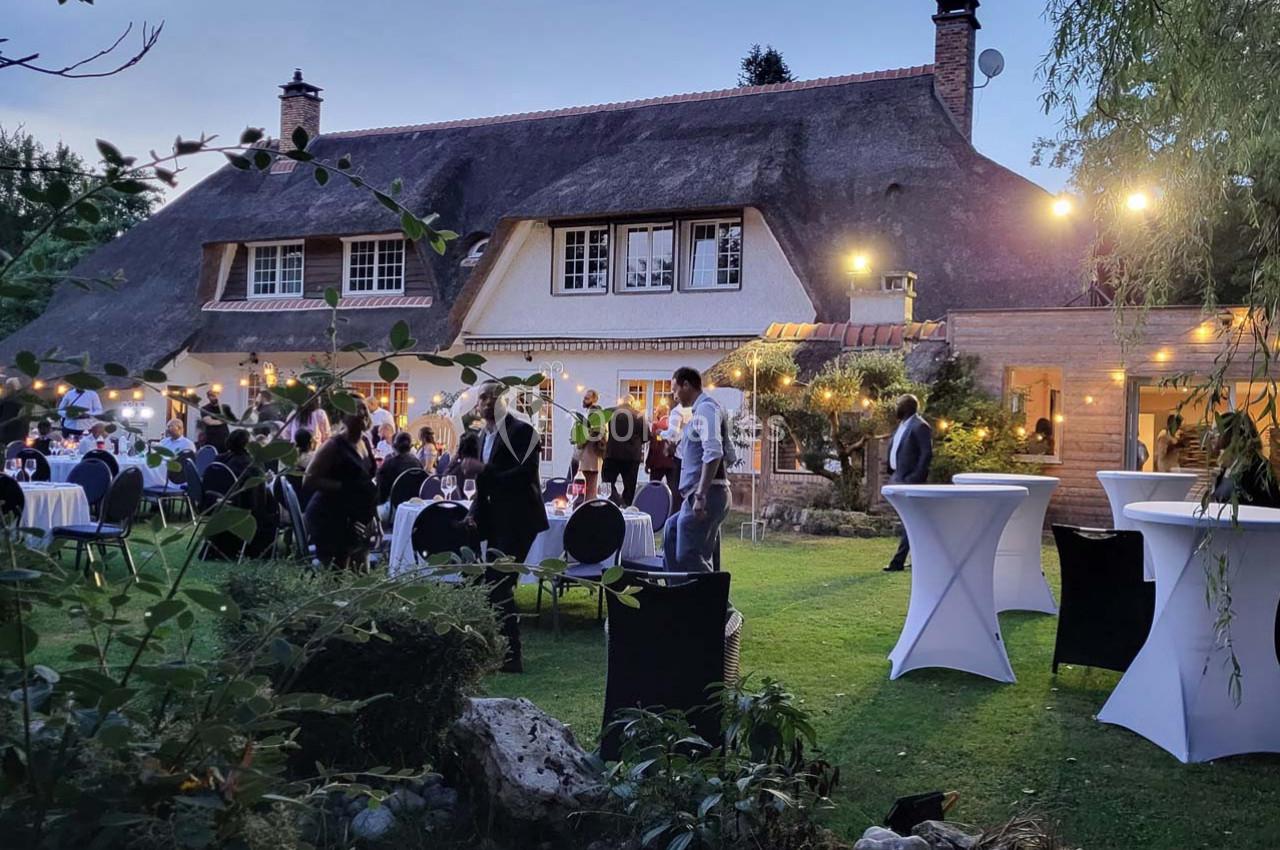 Personnes rassemblées dans un jardin devant une maison au toit de chaume, éclairée par des guirlandes lumineuses.