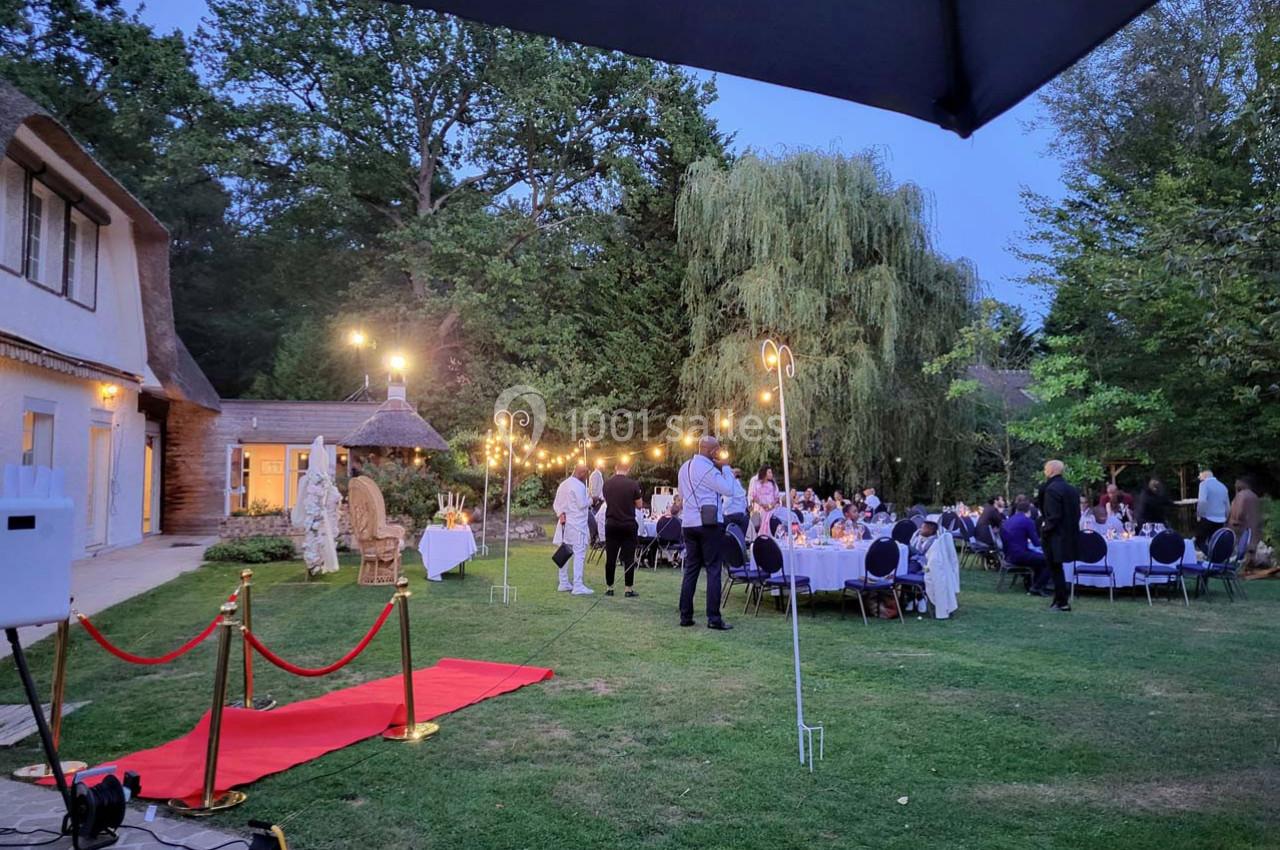 Soirée en plein air avec des invités assis à des tables rondes, éclairée par des guirlandes lumineuses dans un jardin.
