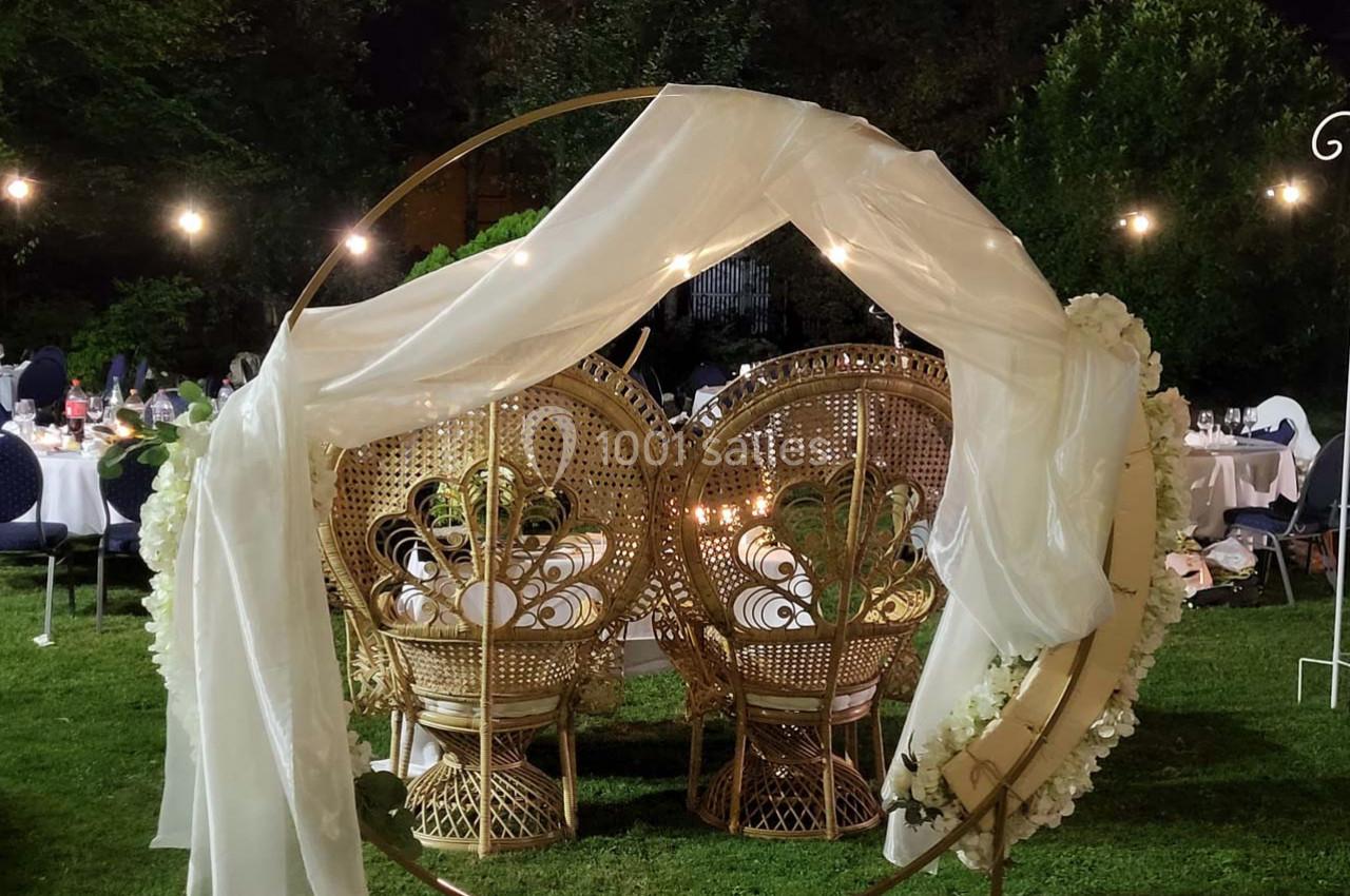 Deux fauteuils en rotin ornés de fleurs et drapés blancs, disposés dans un cercle métallique sur une pelouse en soirée.