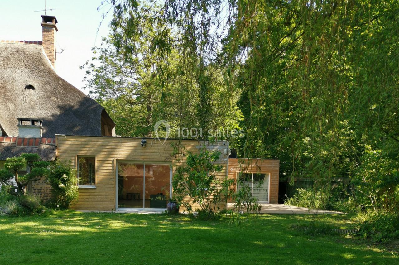 Maison en bois avec grandes baies vitrées, entourée d'arbres et d'un jardin verdoyant sous un ciel dégagé.