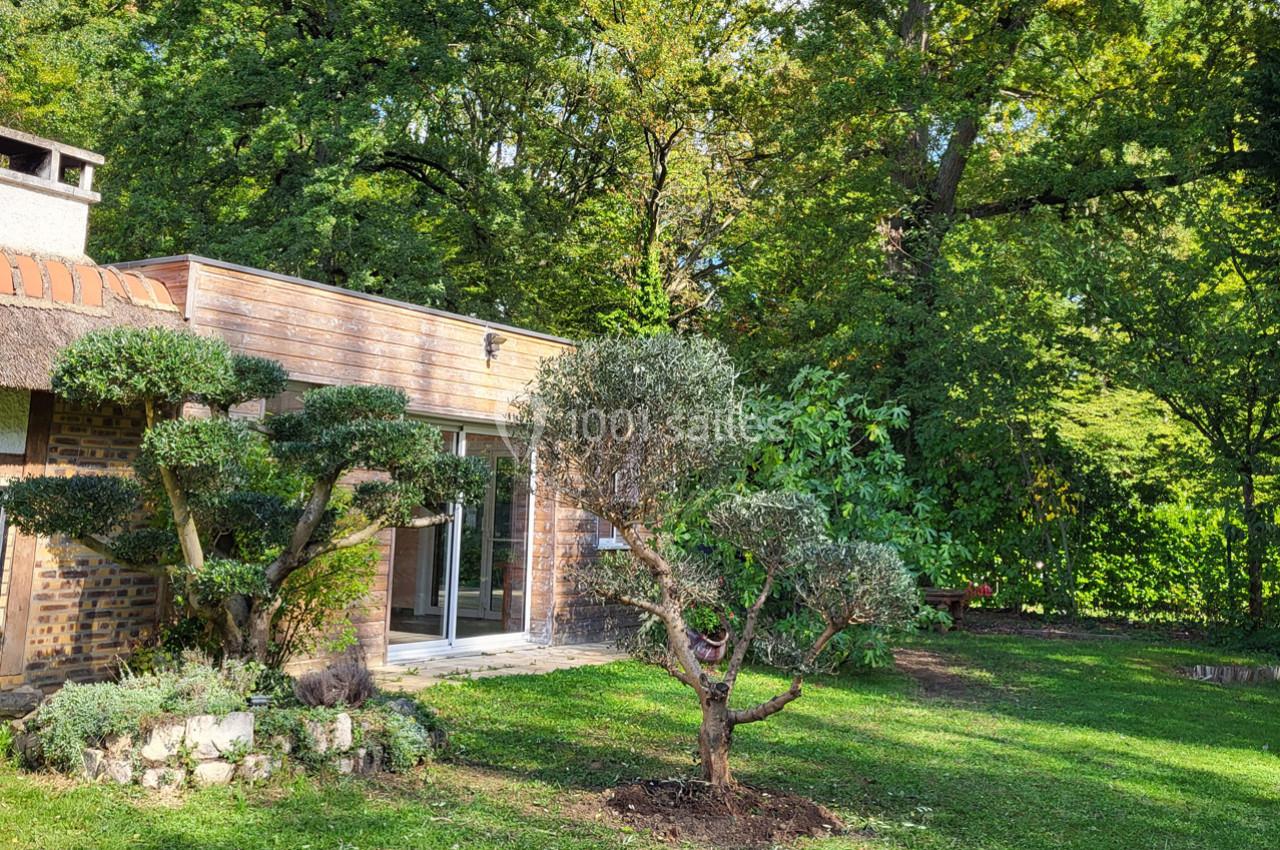 Maison en bois avec de grandes baies vitrées, entourée d'arbres et d'un jardin verdoyant sous un ciel ensoleillé.