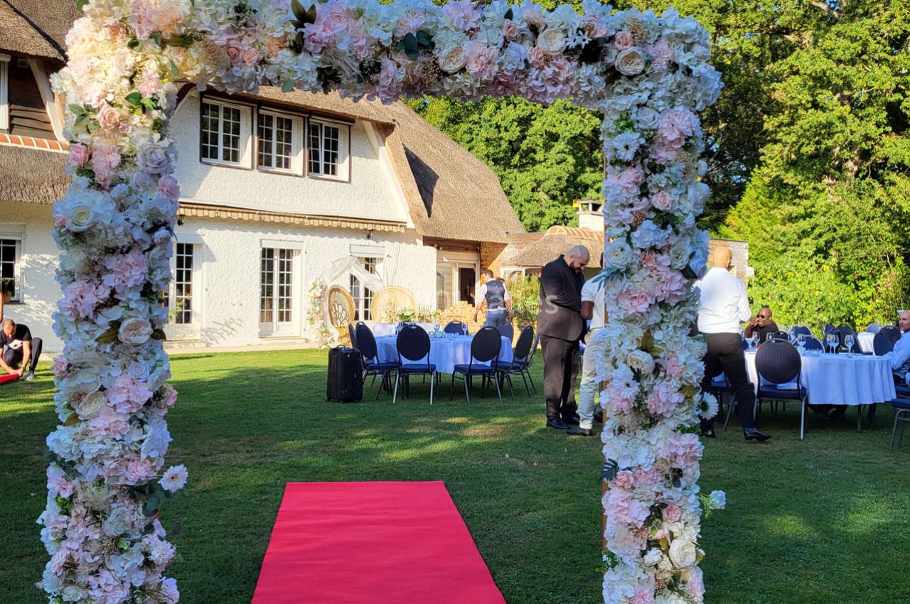 Arche fleurie avec tapis rouge menant à des tables dressées en extérieur devant une maison au toit de chaume.