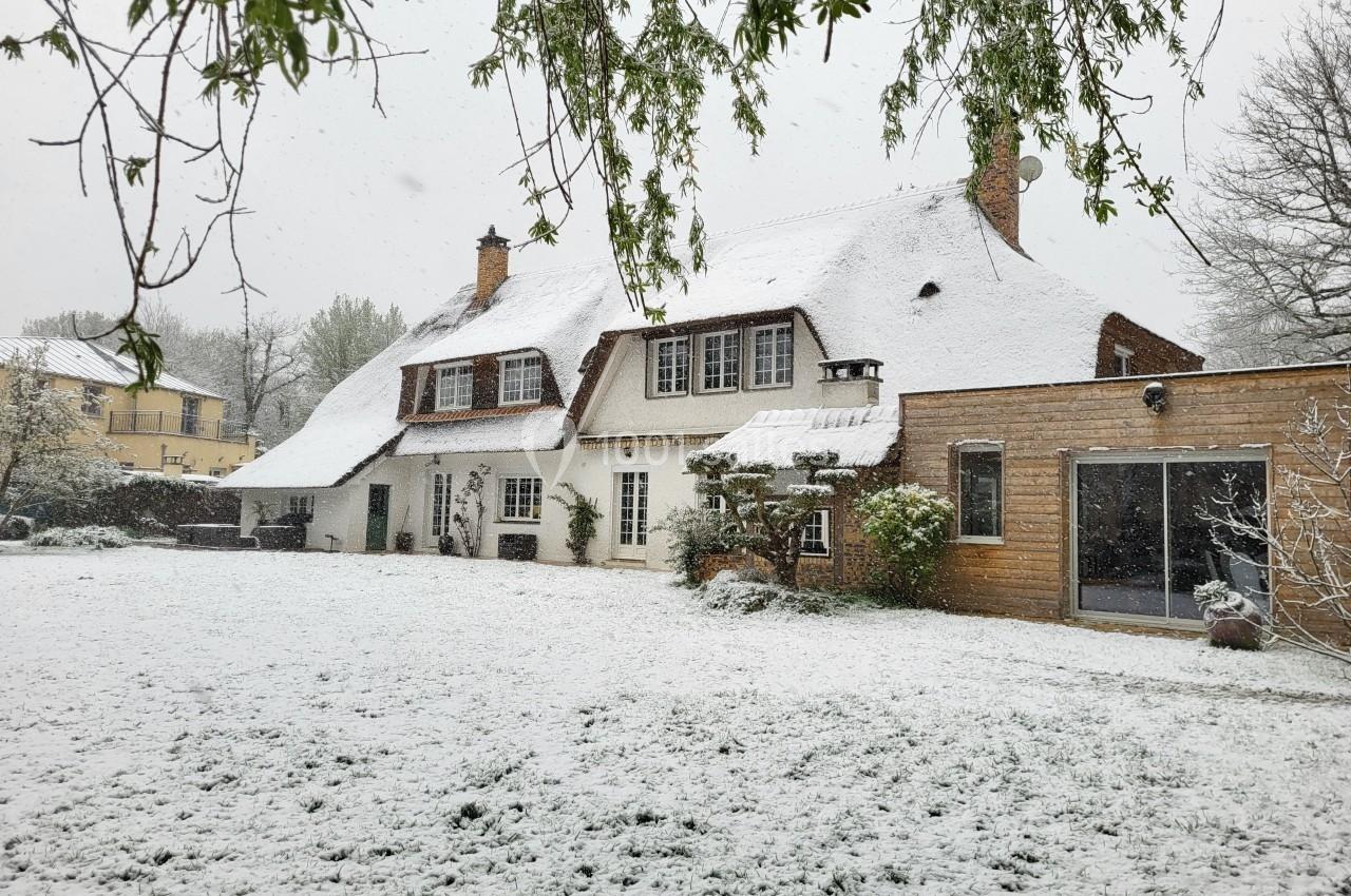 Maison traditionnelle avec toit enneigé, jardin recouvert de neige et arbres en arrière-plan.