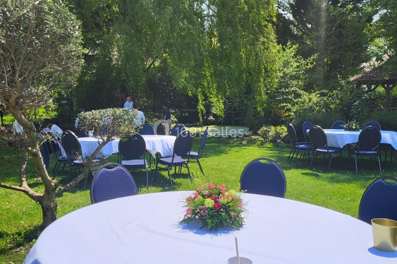 Tables rondes dressées avec nappes blanches et décorations florales dans un jardin verdoyant et ensoleillé.