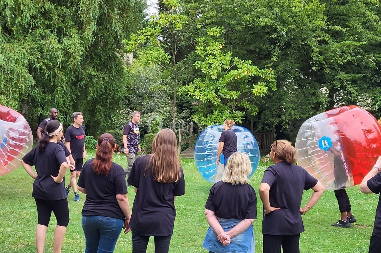 Des personnes en tenue décontractée observent un groupe jouant au bubble football dans un parc verdoyant.