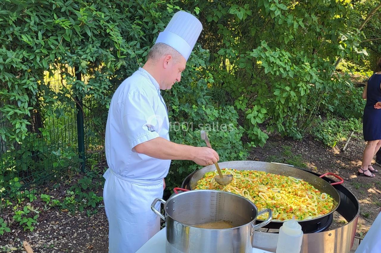Un chef en tenue blanche prépare une grande paella en extérieur, entouré de verdure.