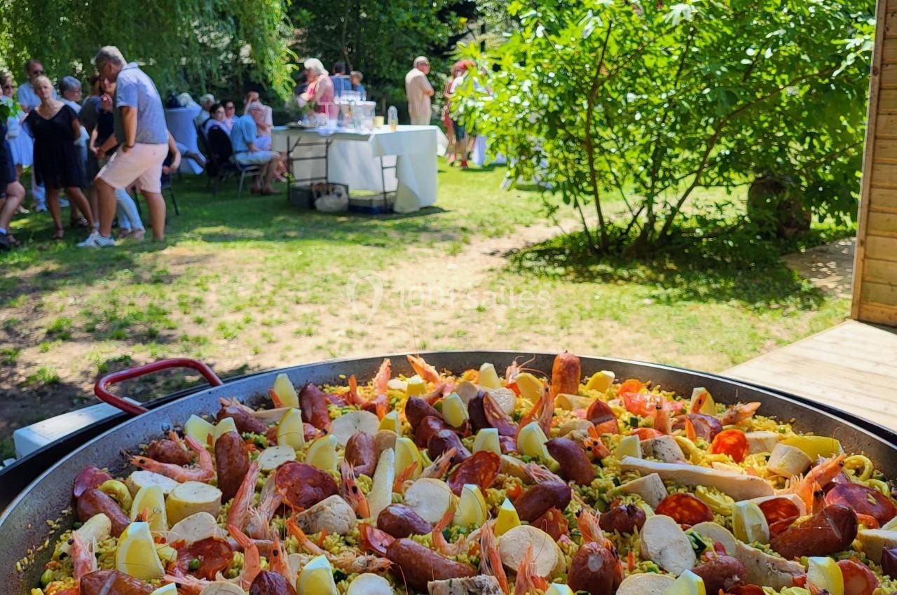 Grande poêle de paella garnie de fruits de mer, saucisses et quartiers de citron, dans un jardin avec des convives en…