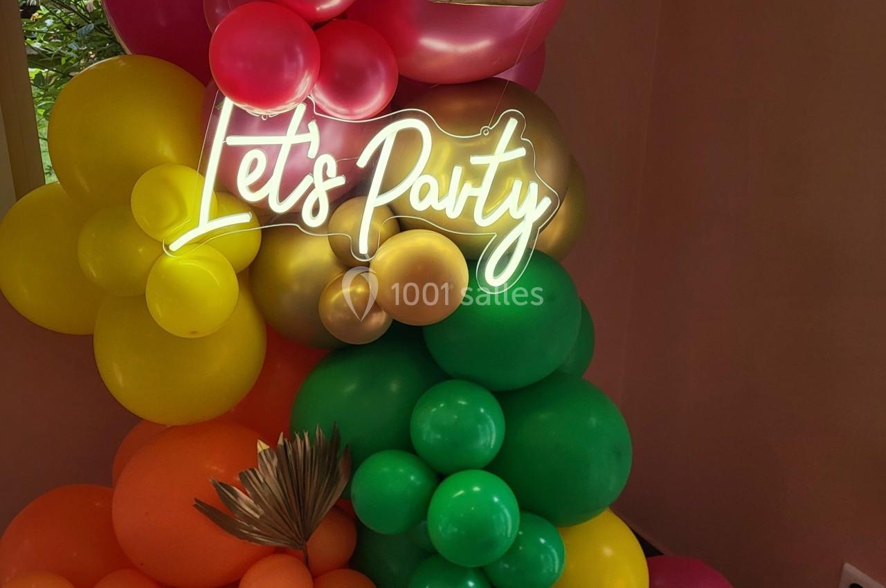 Décoration festive avec des ballons colorés et un néon ’Let's Party’ sur un fond intérieur.