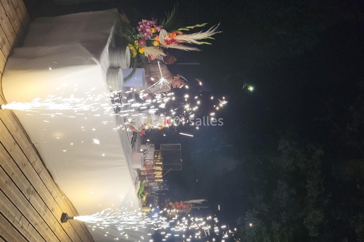 Des étincelles jaillissent devant une table décorée avec des fleurs et des plats, sur une terrasse en soirée.
