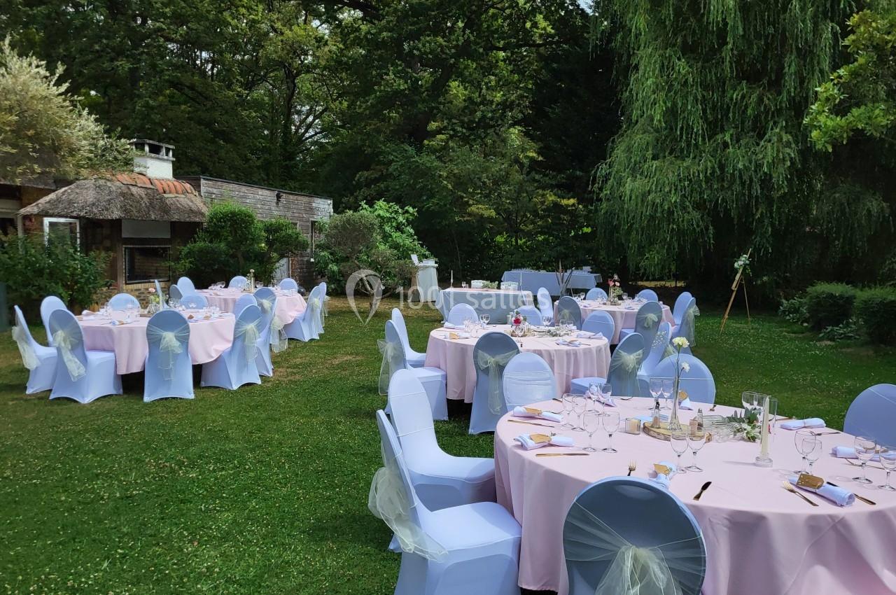 Tables rondes dressées avec nappes roses et chaises bleues, disposées dans un jardin verdoyant sous des arbres.