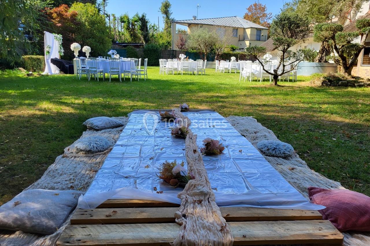 Table basse en bois décorée pour un événement en plein air, entourée de coussins, avec des tables dressées en arrière-plan.