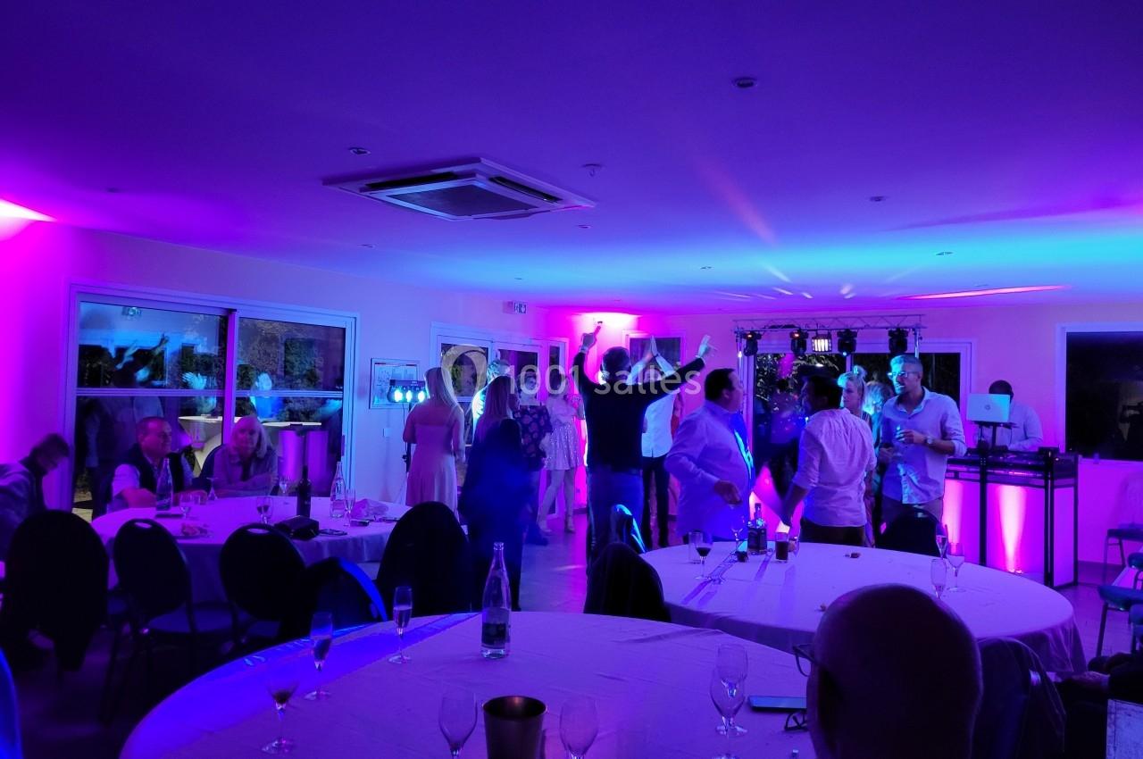 Personnes dansant et discutant dans une salle éclairée par des lumières colorées, tables avec verres au premier plan.