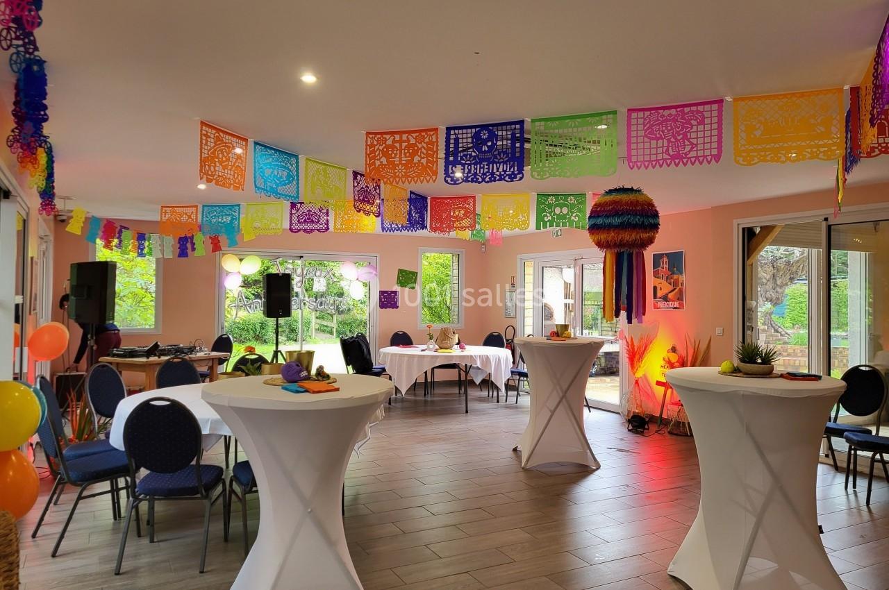 Salle décorée avec des guirlandes colorées, des tables hautes et des chaises, prête pour un événement festif.