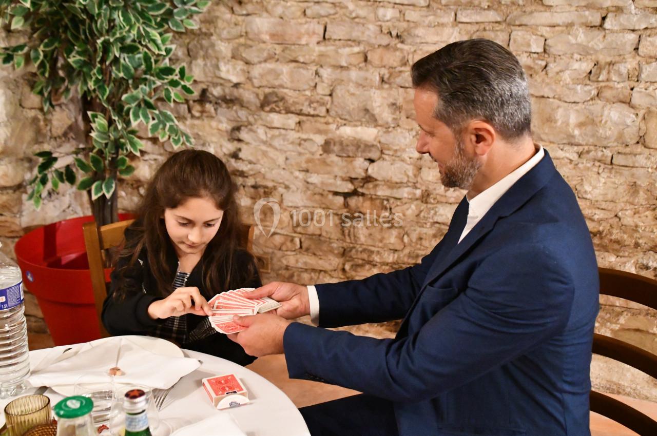 Un homme en costume montre un jeu de cartes à une jeune fille concentrée, assise à une table dans un décor en briques.