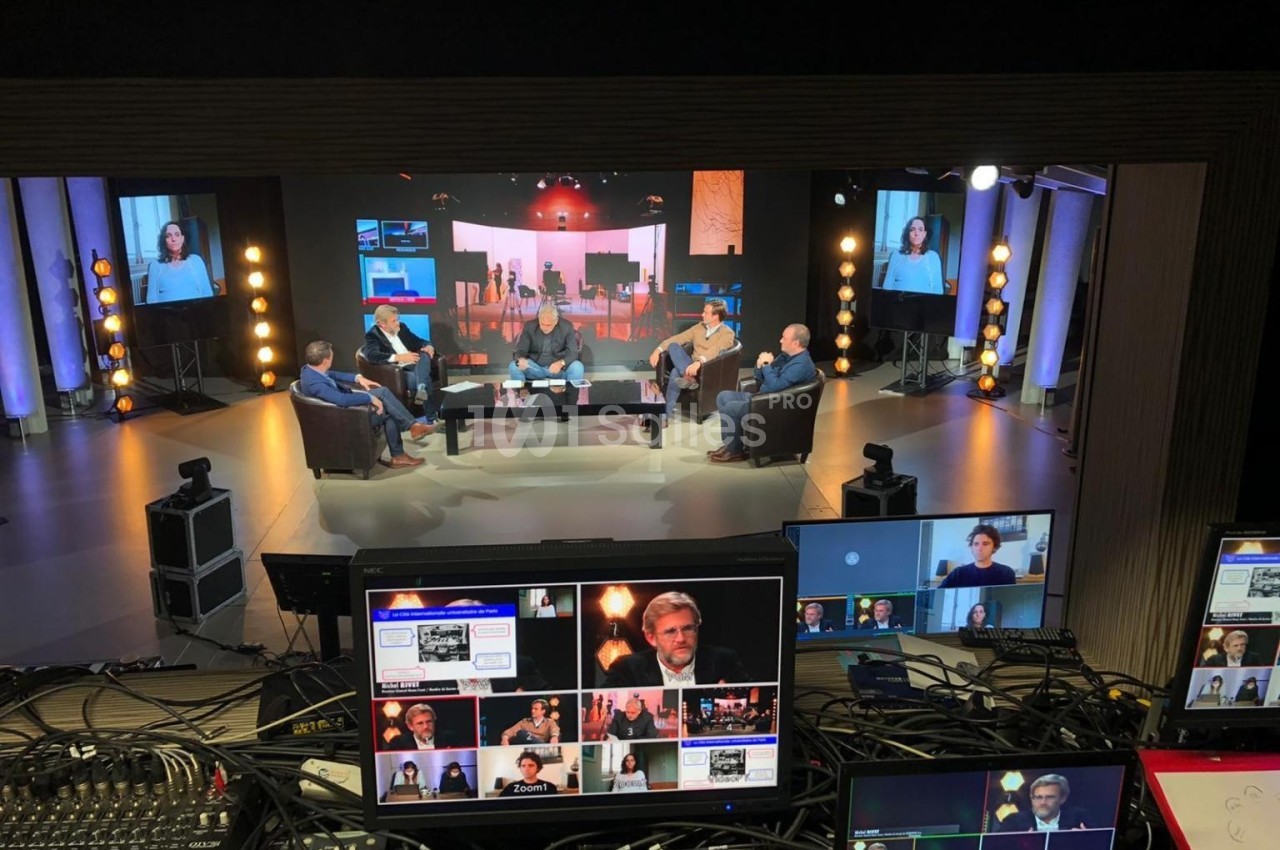 Plateau de tournage avec plusieurs personnes discutant autour d'une table, vu depuis une régie technique.