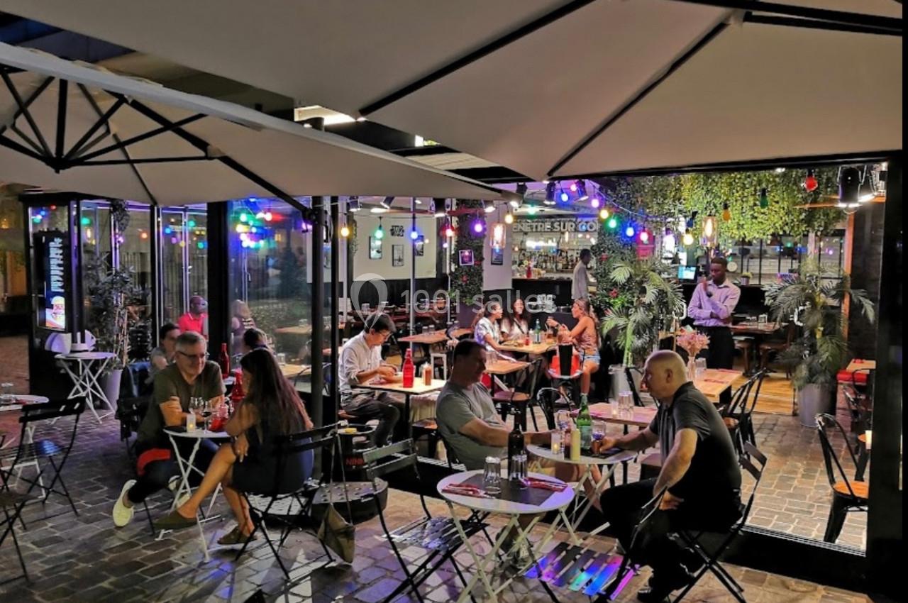 Terrasse d'un restaurant en soirée, avec des clients attablés sous des parasols et des lumières colorées suspendues.