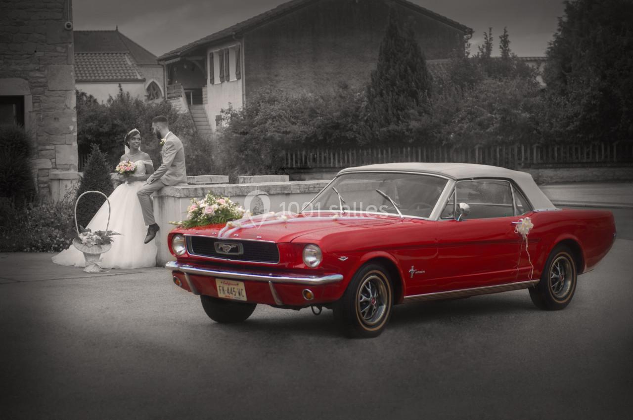 Voiture ancienne rouge avec capote blanche au premier plan, couple de mariés en arrière-plan devant un bâtiment.
