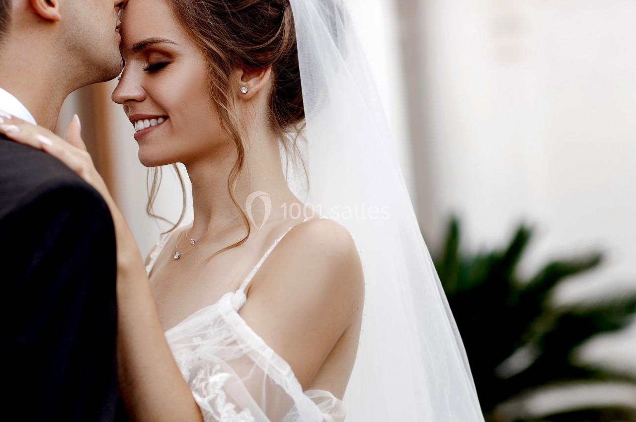 Une mariée souriante en robe blanche et voile, embrassée sur le front par un homme en costume.