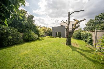 Jardin avec pelouse, arbre taillé, haies verdoyantes et maison moderne en arrière-plan sous un ciel partiellement nuageux.