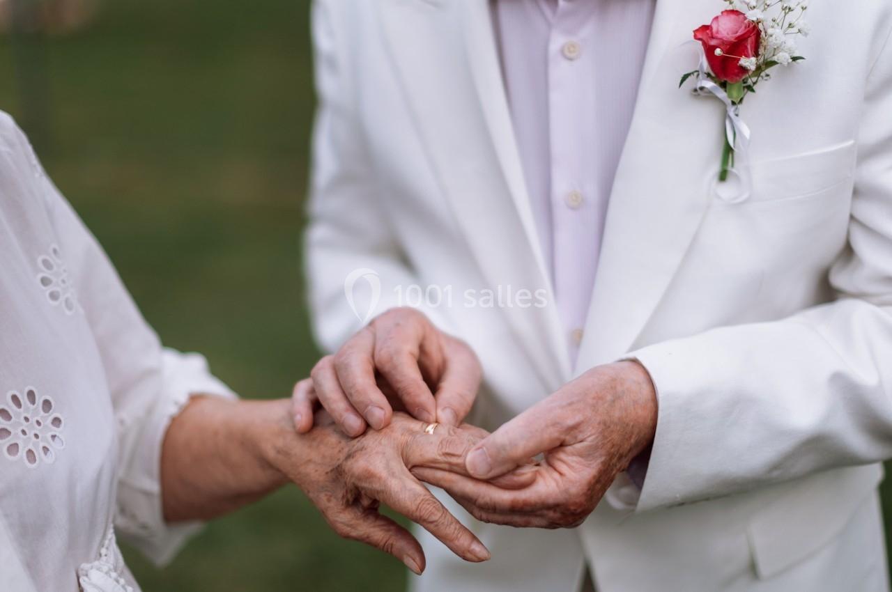 Un homme en costume blanc glisse une bague au doigt d'une femme, dans un cadre extérieur verdoyant.